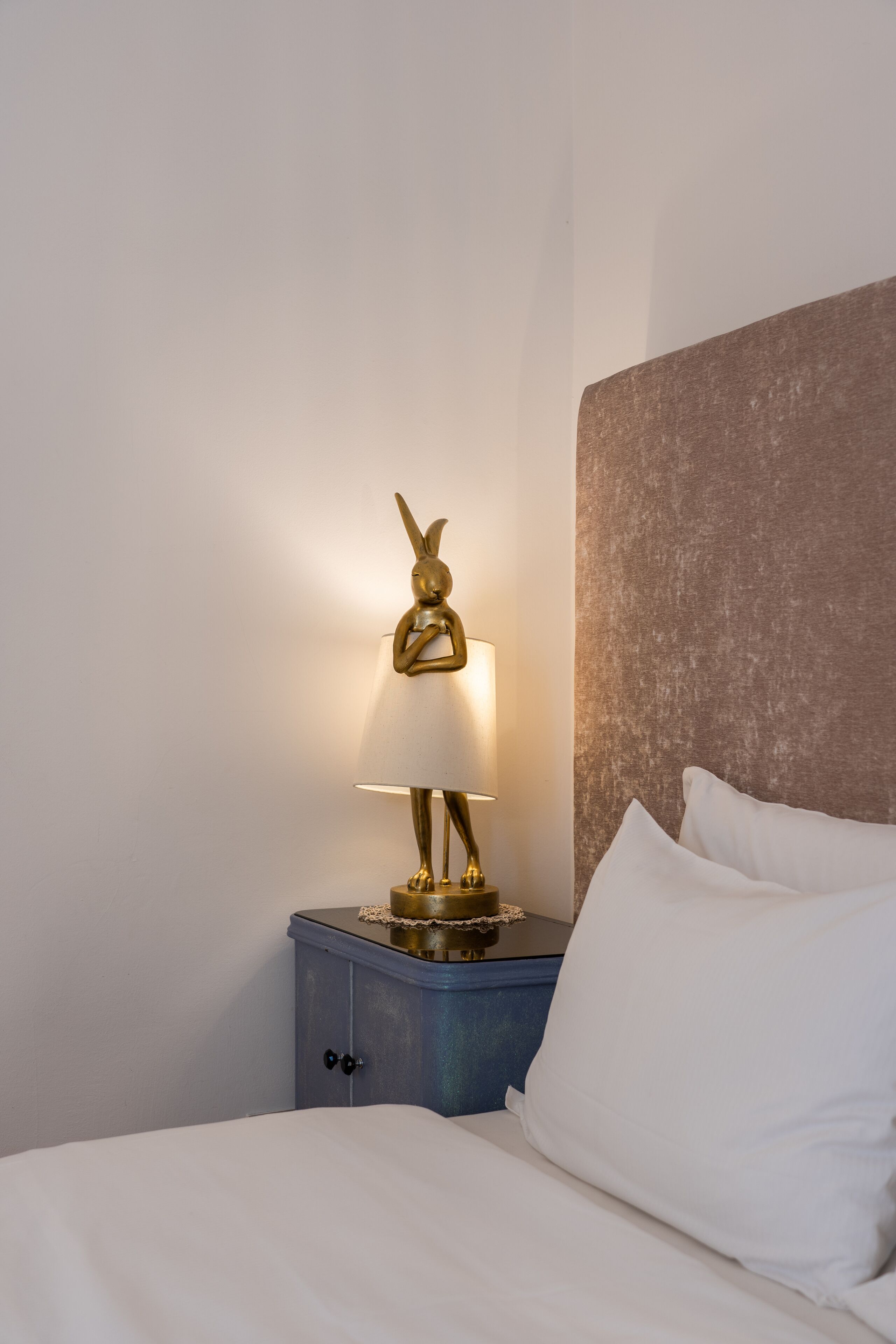 A neatly made bed with white linens, a small bedside table with a lamp featuring a golden rabbit sculpture, and a textured brown headboard.