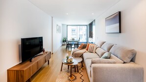 A modern living room with a flat-screen TV on a wooden console, a beige sofa, a coffee table, and a dining area in the background. - Camões Downtown with Parking (Porto)