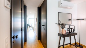 A modern hallway with a wooden floor, a black door, a white wall with an air conditioning unit, a desk with a vase, and a floor lamp. - Camões Downtown with Parking (Porto)