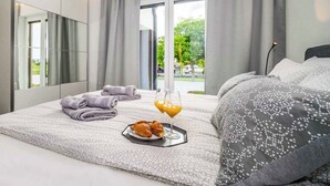 Ein modernes Hotelzimmer mit einem großen Bett, einem Nachttisch mit einem Tablett voller Croissants und Orangensaft sowie einem Blick ins Freie durch ein großes Fenster.