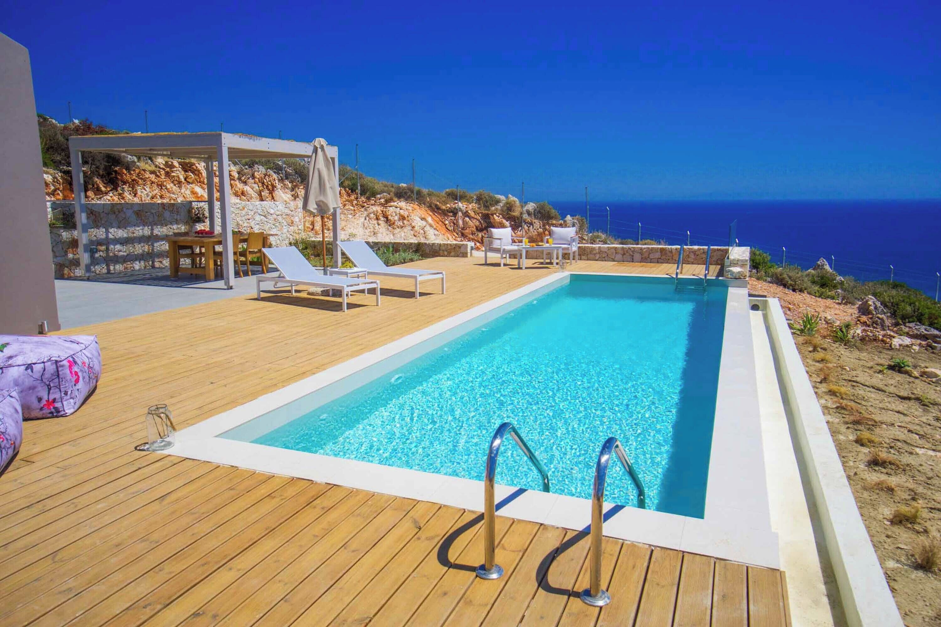 A rectangular swimming pool with a wooden deck, surrounded by lounge chairs and a pergola, overlooking a rocky coastline.