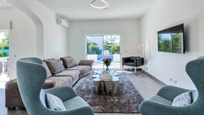 Ein modernes Wohnzimmer mit grauem Sofa, zwei Sesseln, einem Couchtisch und einem an der Wand befestigten Flachbildfernseher.