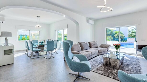 Ein geräumiges Wohnzimmer im modernen Stil, mit einem grauen Sofa, türkisfarbenen Sesseln, einem Essbereich mit einem Glastisch und großen Fenstern mit Blick auf einen Swimmingpool.