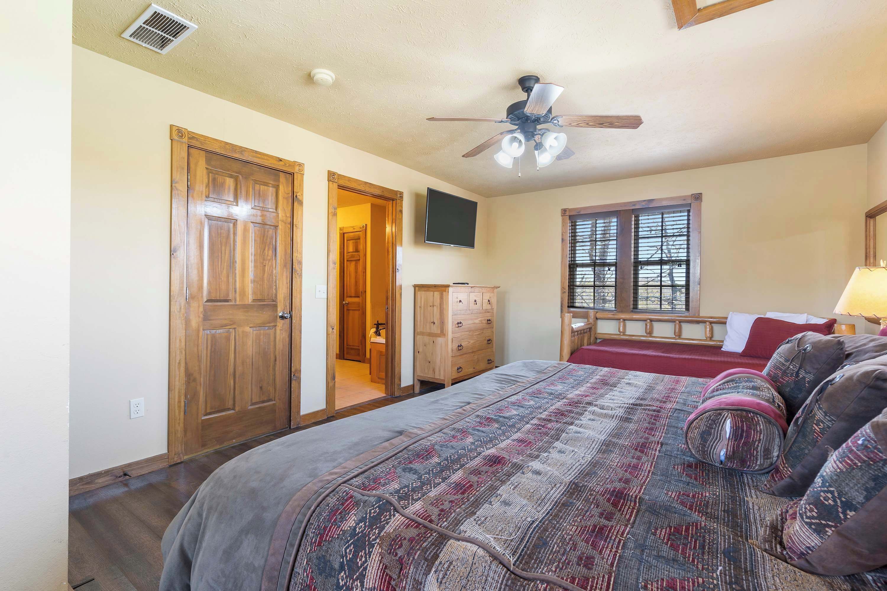 A bedroom with a large bed, a ceiling fan, a wooden wardrobe, a dresser, and a window with blinds.