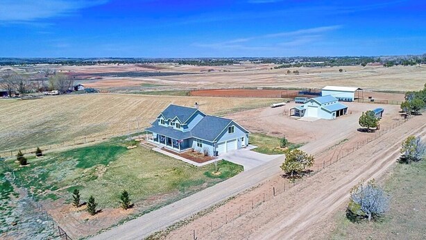 Une grande maison avec un garage, entourée de champs ouverts et bordée d’un chemin de terre.