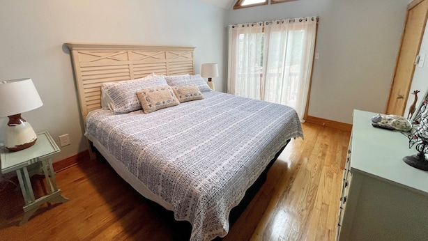 A bedroom with a wooden bed, a nightstand with a lamp, a window with curtains, and a wooden floor.