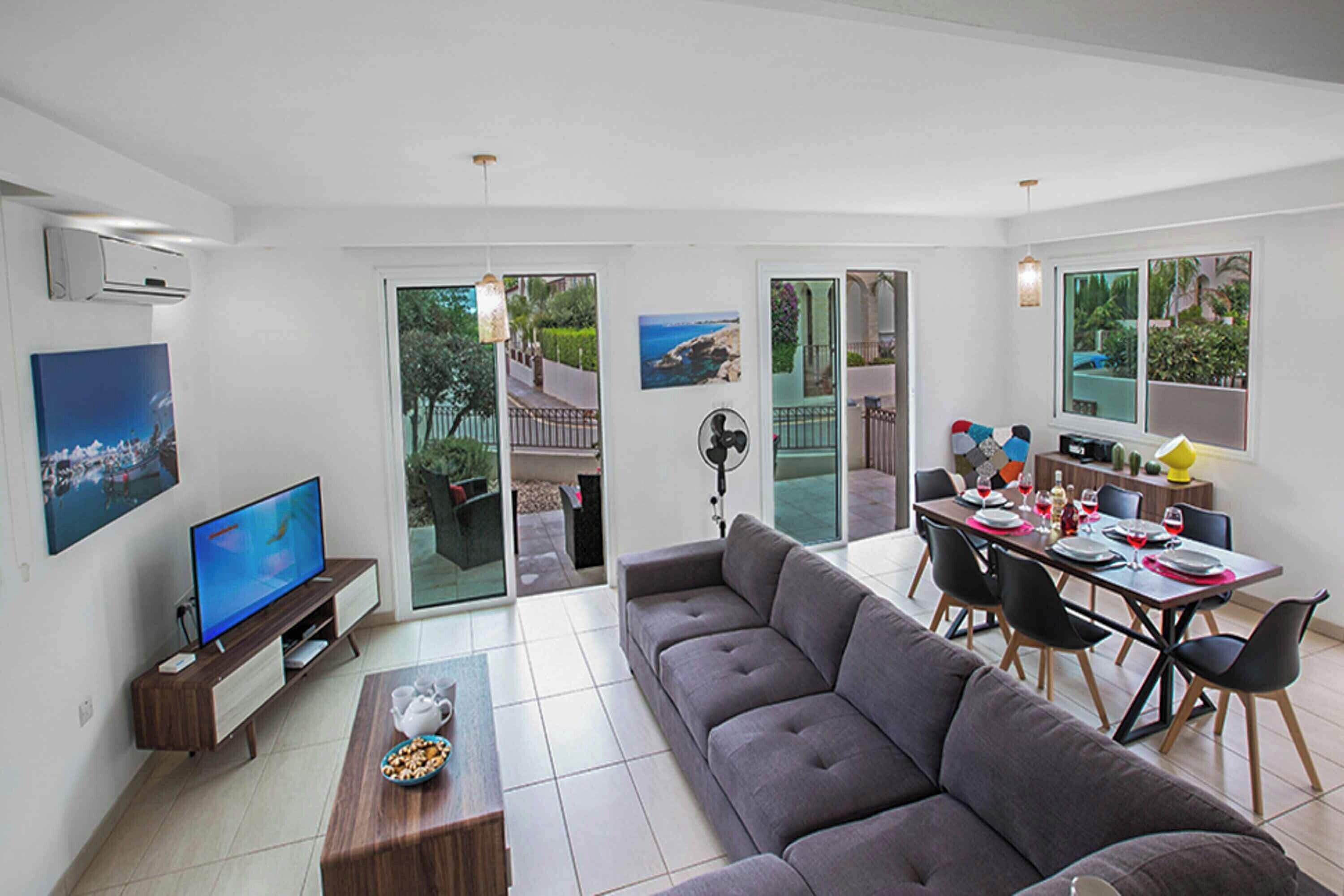 A modern living room with a grey sofa, a dining table set for four, and a television on a wooden stand.