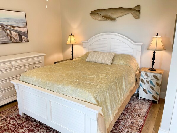 Un dormitorio con cama blanca, dos lámparas de noche, una mesita de noche con motivos de peces, una alfombra estampada y un cuadro con una playa.