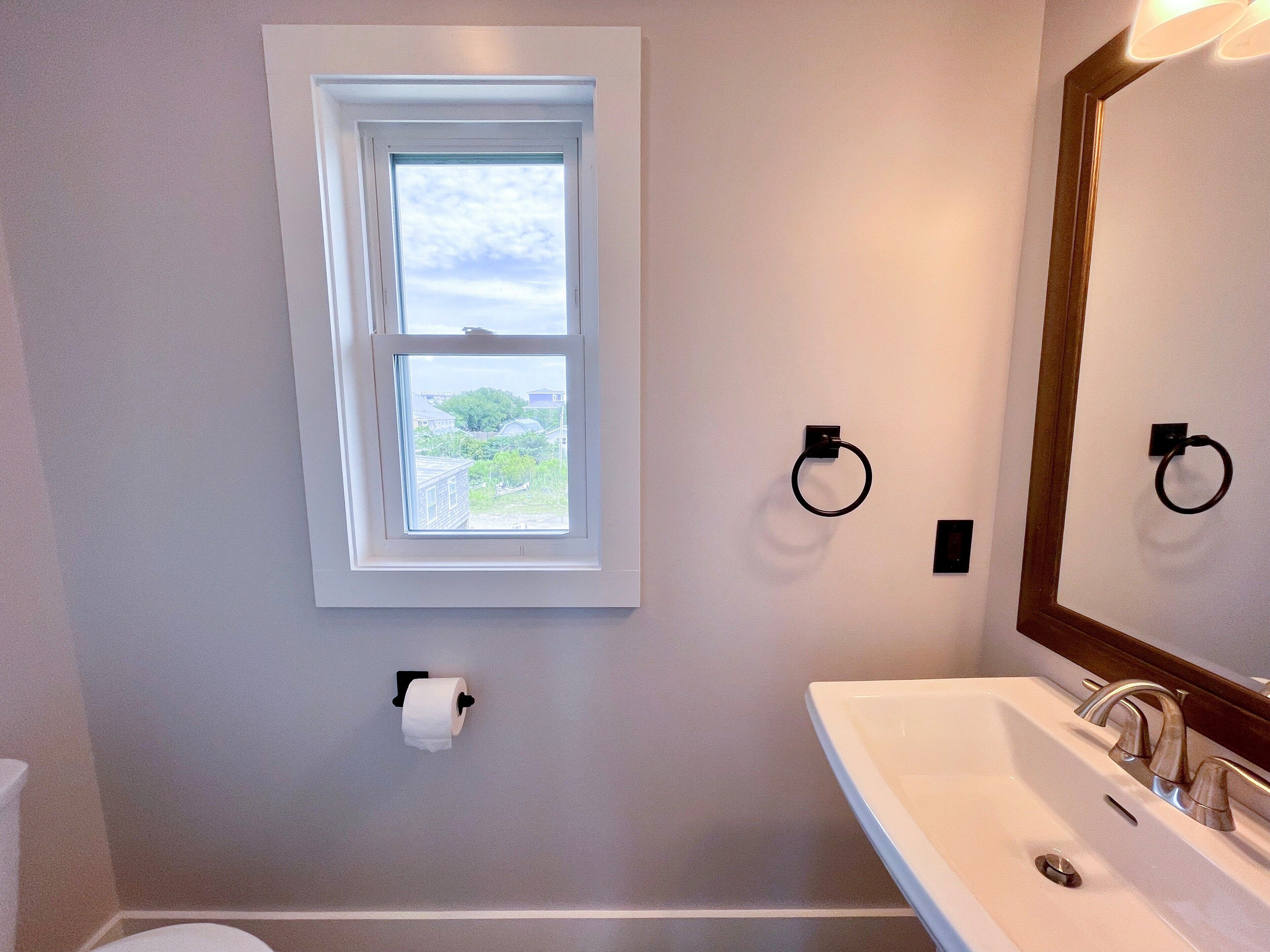A bathroom with a window, a toilet paper holder, a sink with a mirror, and towel rings.