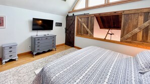 Un dormitorio con techo inclinado, ventilador de techo, televisor, cómoda y ventana con marco de madera.