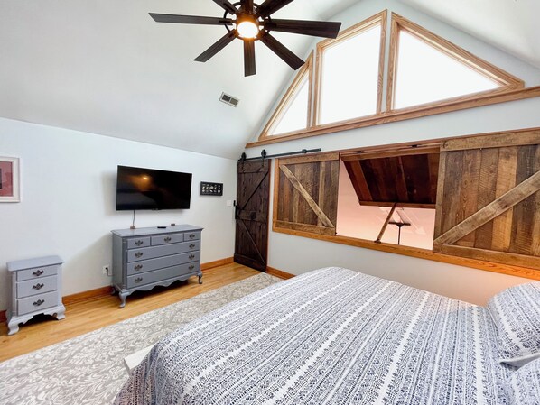 Een slaapkamer met schuine kap, een plafondventilator, een televisie, een dressoir en een raam met houten kader.
