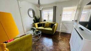 Ein Wohnzimmer mit Fischgrätparkettboden, einem gelben Sofa, einem grünen Sessel, einem Glastisch und einem Fernseher auf einem weißen Schrank.