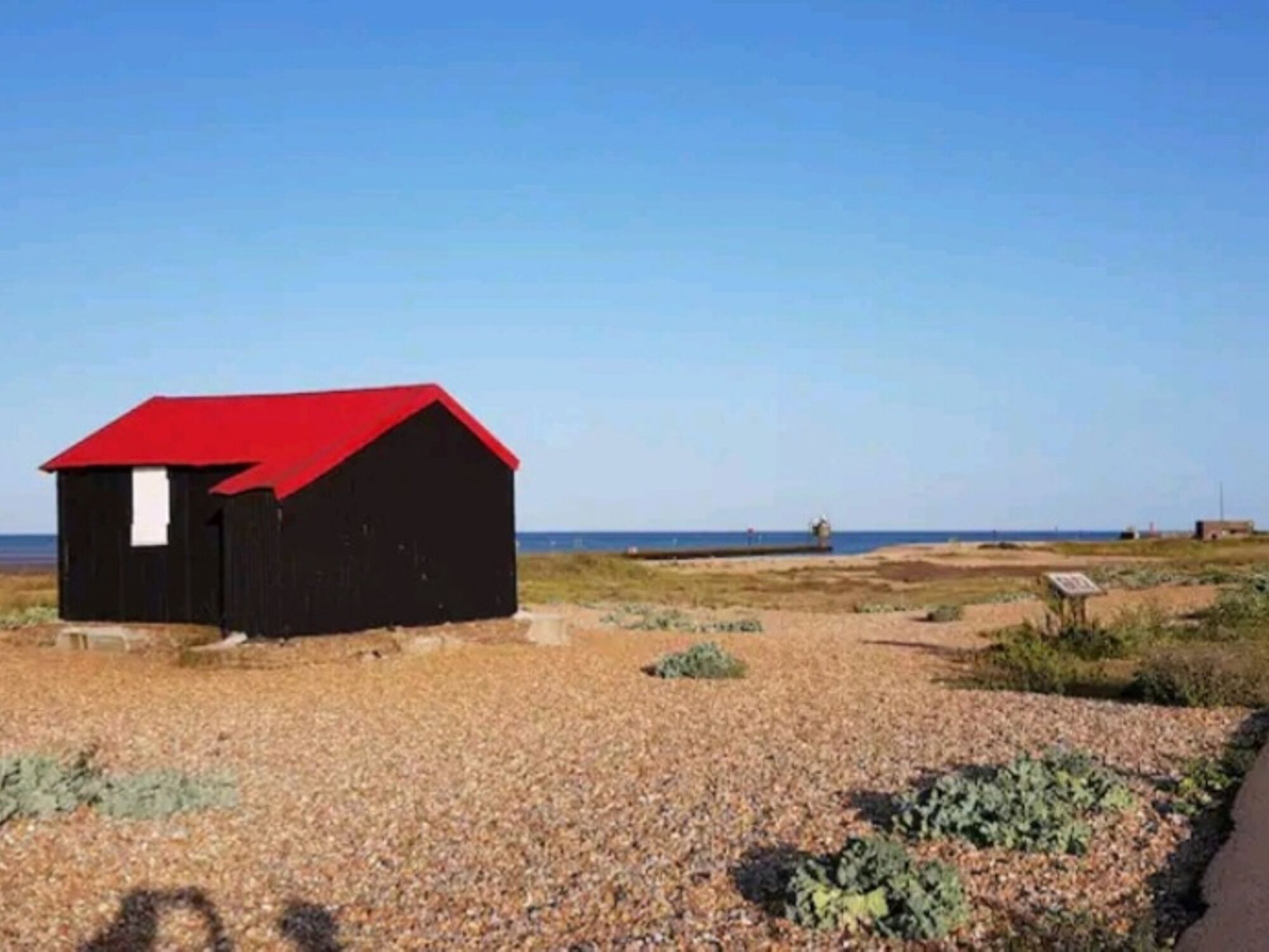 Un petit bâtiment noir avec un toit rouge, situé sur un terrain recouvert de gravier où la végétation est clairsemée.