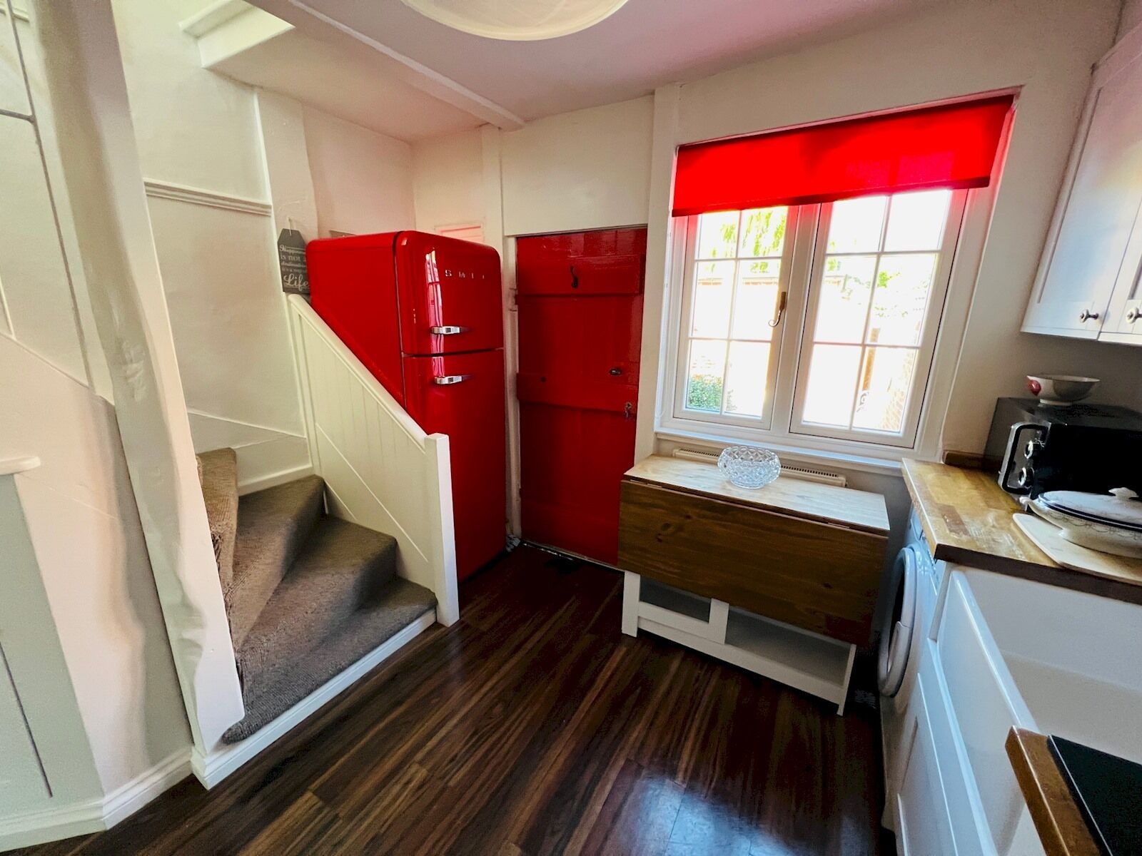 A compact kitchen with a red refrigerator, a wooden countertop, and a window with a red blind.