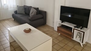 Una sala de estar con un sofá gris, una mesa de centro blanca, un televisor de pantalla plana sobre un soporte de madera y un equipo de aire acondicionado en la pared.