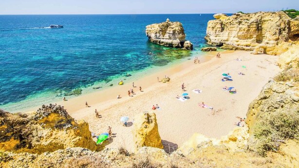 Una playa con agua cristalina, formaciones rocosas y gente tomando el sol.