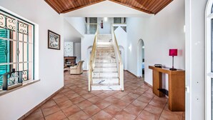 Un hall d’entrée spacieux, avec un escalier en bois menant à l’étage supérieur, un sol carrelé et une petite table avec une lampe sur le côté droit.