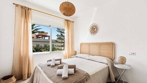 A bedroom with a large bed, a window with curtains, a small round table with a lamp, and a wall decoration.