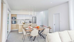 A modern kitchen with white cabinets, a dining table set for four, and a sofa in the living area. - Vie Bleu 15 - Three Bedroom Villa, Sleeps 6 (Πρωταράς)