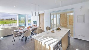 A modern kitchen with a large island, dining area with chairs, and a view of a green landscape. - Vie Bleu 15 - Three Bedroom Villa, Sleeps 6 (Πρωταράς)