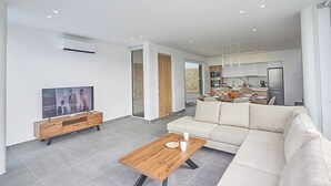 A modern living room with a flat-screen TV, a wooden coffee table, a beige sofa, and a kitchen area visible through an open door. - Vie Bleu 15 - Three Bedroom Villa, Sleeps 6 (Πρωταράς)