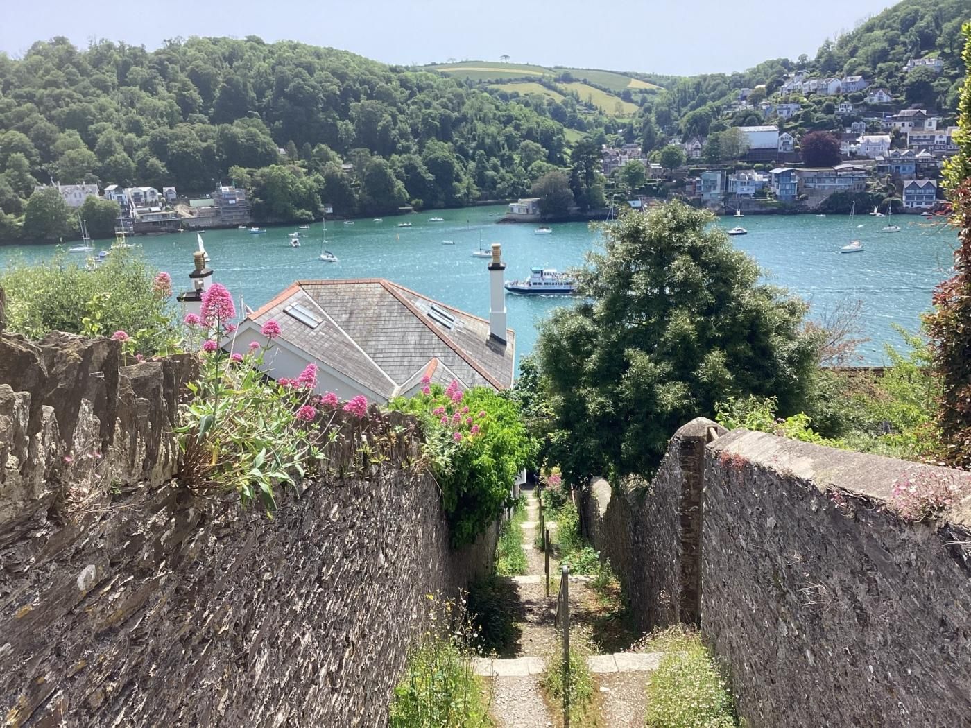 A coastal town with a stone wall, a pathway leading to a building with a sloped roof, and boats on the water.