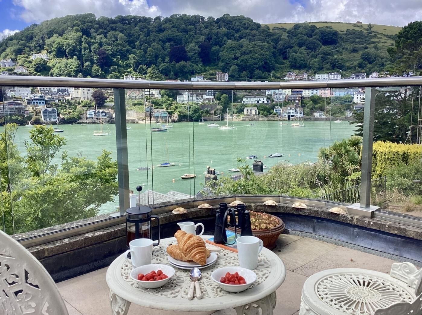 A balcony with a table set for breakfast, overlooking a harbor with boats and houses in the background.