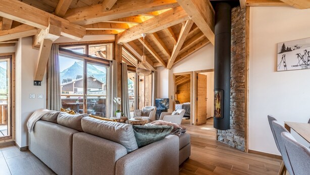 A spacious living room with a large sofa, wooden beams, a stone fireplace, and large windows offering mountain views.