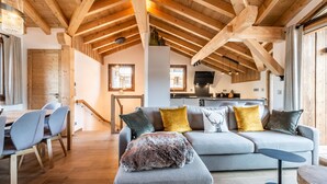 A spacious living area with a high wooden ceiling, a gray sofa with multiple cushions, a dining table with chairs, and a small round table.
