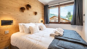 A bedroom with a wooden wall, a bed with white bedding, a window with curtains, and a view of a building outside.