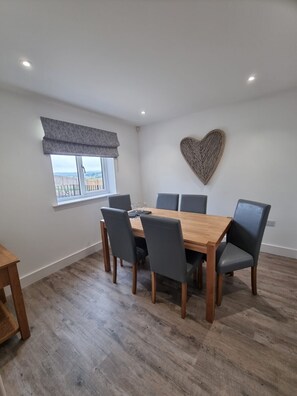 A dining area with a wooden table and six chairs, a window with a view, and a heart-shaped wall decoration. - Stone built cottage, comfortably sleeps up to 6.<br> <br>Ideal for holiday makers who love to explore! (Morpeth)