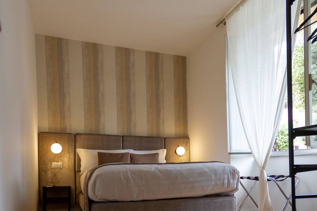 A bedroom with a bed, wooden flooring, a headboard with built-in lighting, and a window with curtains.