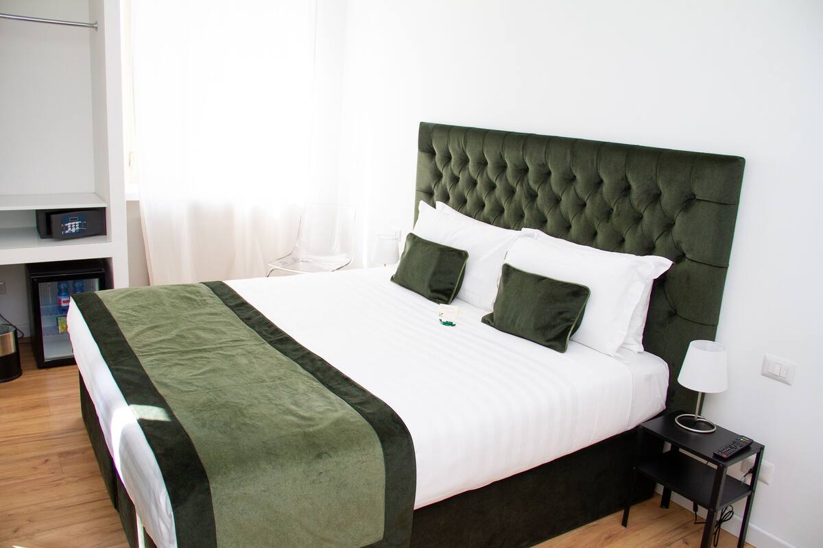 A neatly made bed with a green headboard, white bedding, and green pillows. There is a bedside table with a lamp and a remote control.