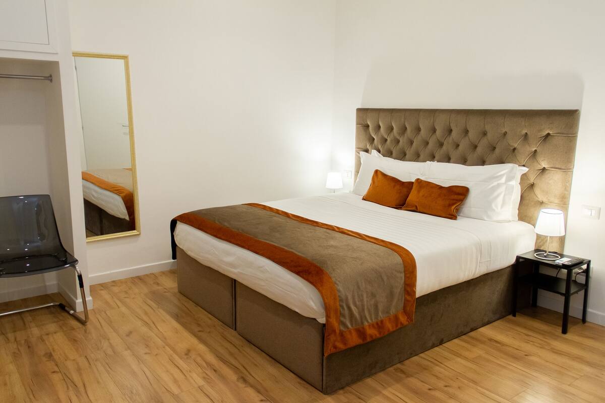 A neatly arranged bedroom with a large bed, a bedside table with a lamp, a chair, and a mirror on the wall.