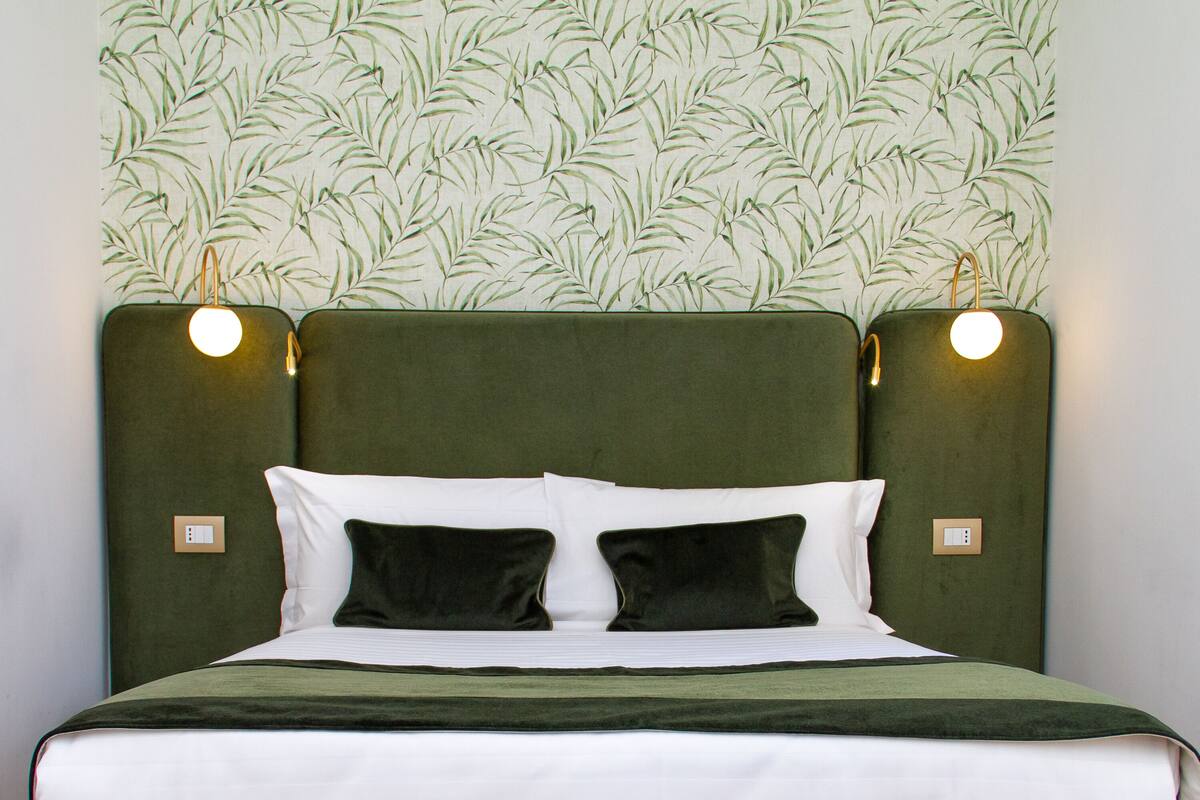 A neatly made bed with a green headboard, two bedside lamps, and a leaf-patterned wallpaper.