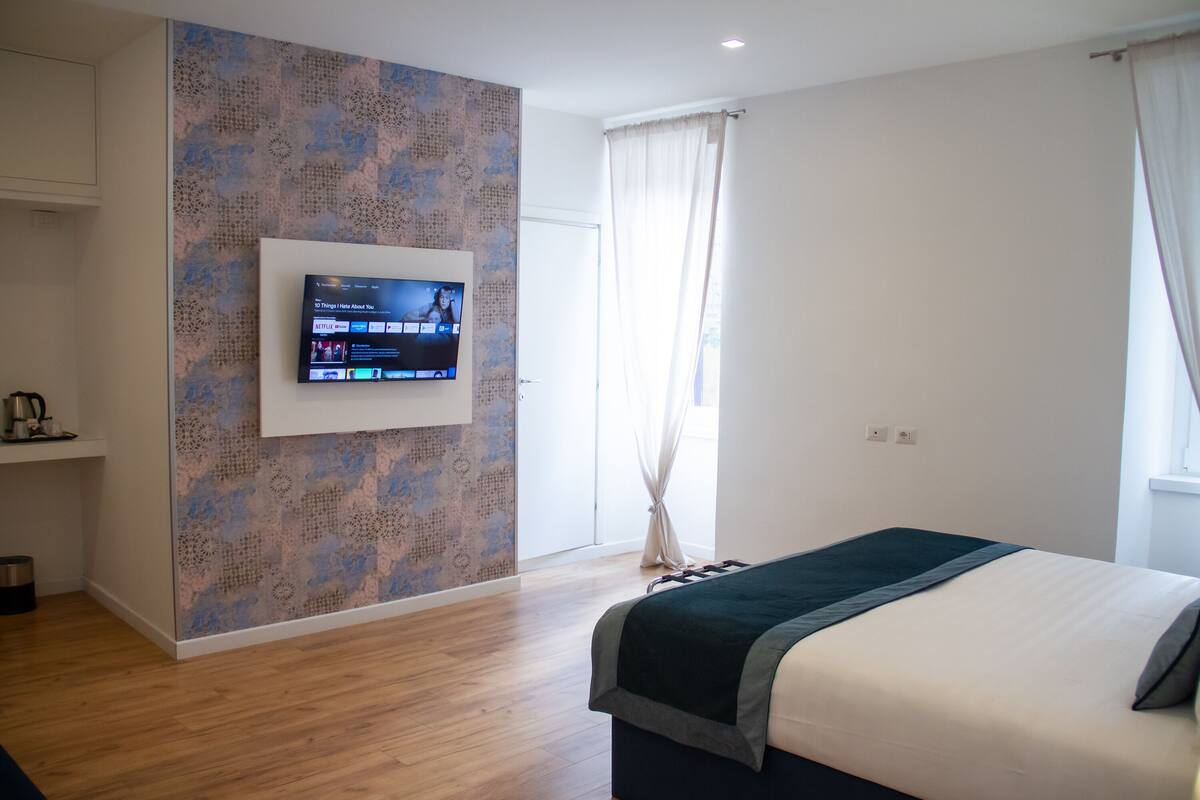 A modern hotel room with a large flat-screen TV mounted on the wall, a bed with a dark headboard, and a wooden floor.