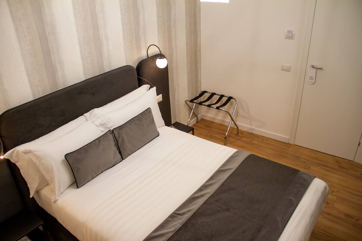 A neatly made bed with white and gray bedding, a bedside lamp, and a small stool.