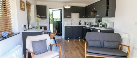 Une cuisine moderne avec des meubles de couleur sombre, un plan de travail blanc et un four à micro-ondes. Le salon est équipé de deux canapés gris et d’une chaise en bois.