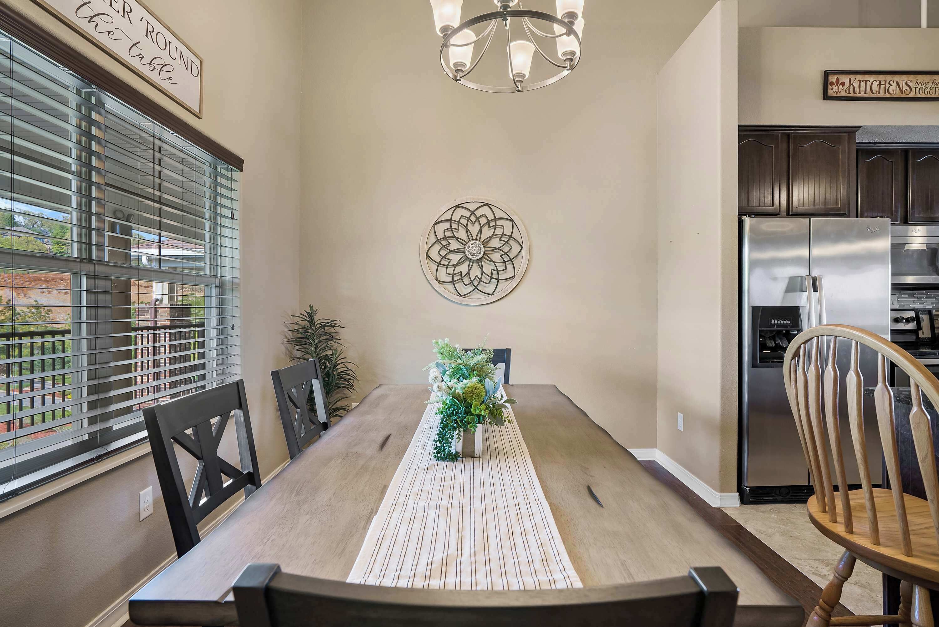 A dining area with a wooden table, chairs, a chandelier, a decorative wall piece, and a refrigerator.