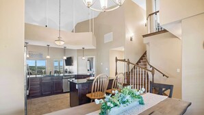 A spacious dining area with a wooden table, chairs, and a floral centerpiece. A kitchen with dark cabinets and a central island is visible in the background. A wooden staircase with a railing leads to an upper level. - Happy Place Condo - Stunning Table Rock Lake Views (Branson)