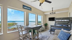 A dining area with a table and chairs, a ceiling fan, a chandelier, a couch with a patterned pillow, and a view of a body of water through large windows. - Happy Place Condo - Stunning Table Rock Lake Views (Branson)