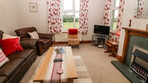 A living room with a brown leather sofa, a wooden coffee table with a striped table runner, a television on a stand, and a fireplace with a mirror above it. - Dinas Farmhouse Annex (Caernarfon)