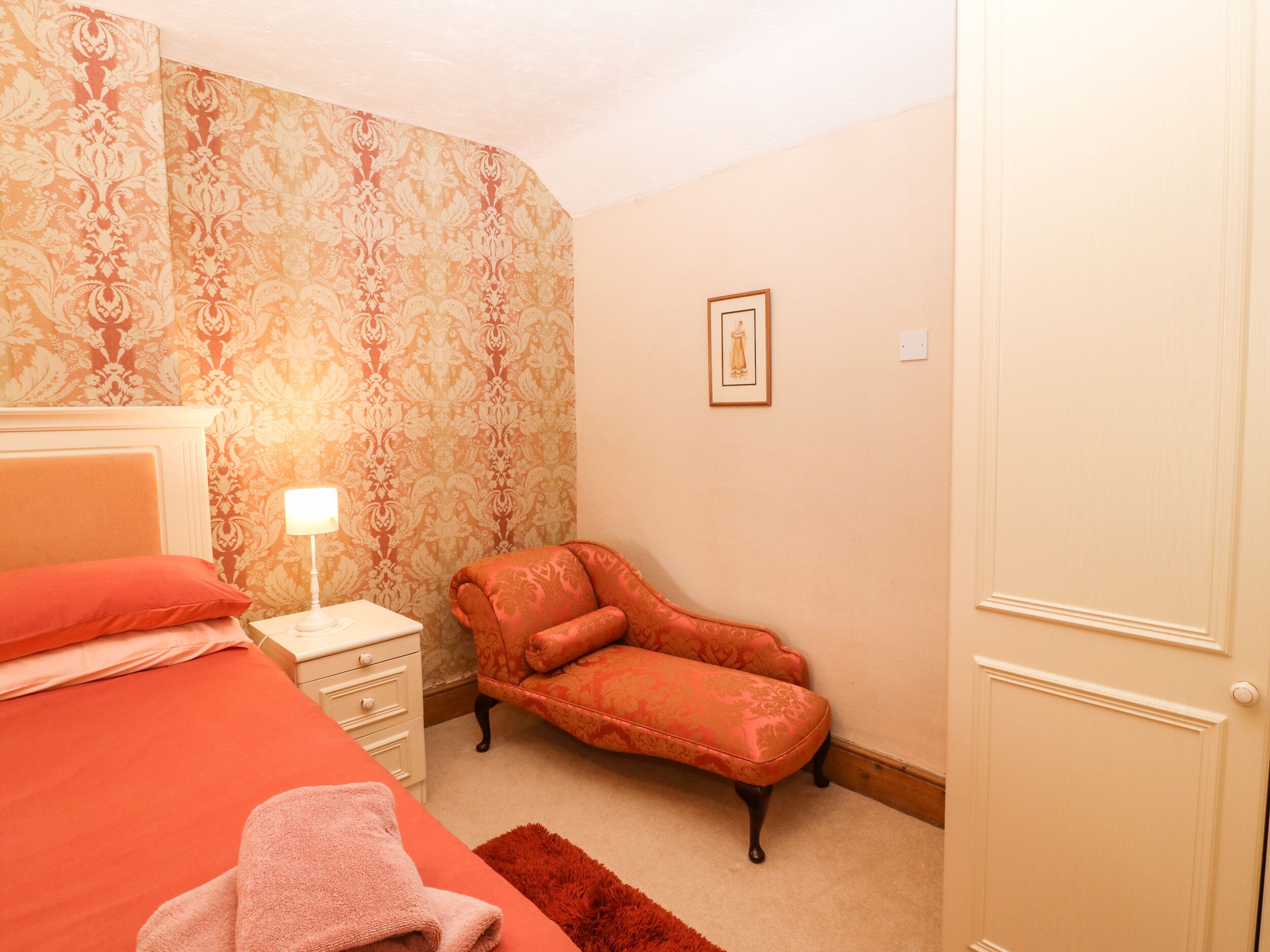 Un dormitorio con cama, mesita de noche, silla, puerta y un cuadro en la pared.
