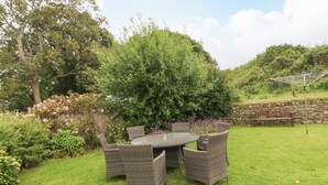 A garden with a round table and wicker chairs, surrounded by lush greenery and flowering plants. - Dinas Farmhouse Annex (Caernarfon)