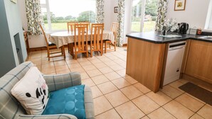A cozy living room with a sofa, a dining table with chairs, and a kitchen area with a dishwasher and a kettle. - Dinas Farmhouse Annex (Caernarfon)