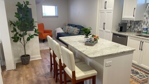 A modern kitchen with a central island, wooden chairs, a potted plant, and a dining area with a sofa and orange chair.