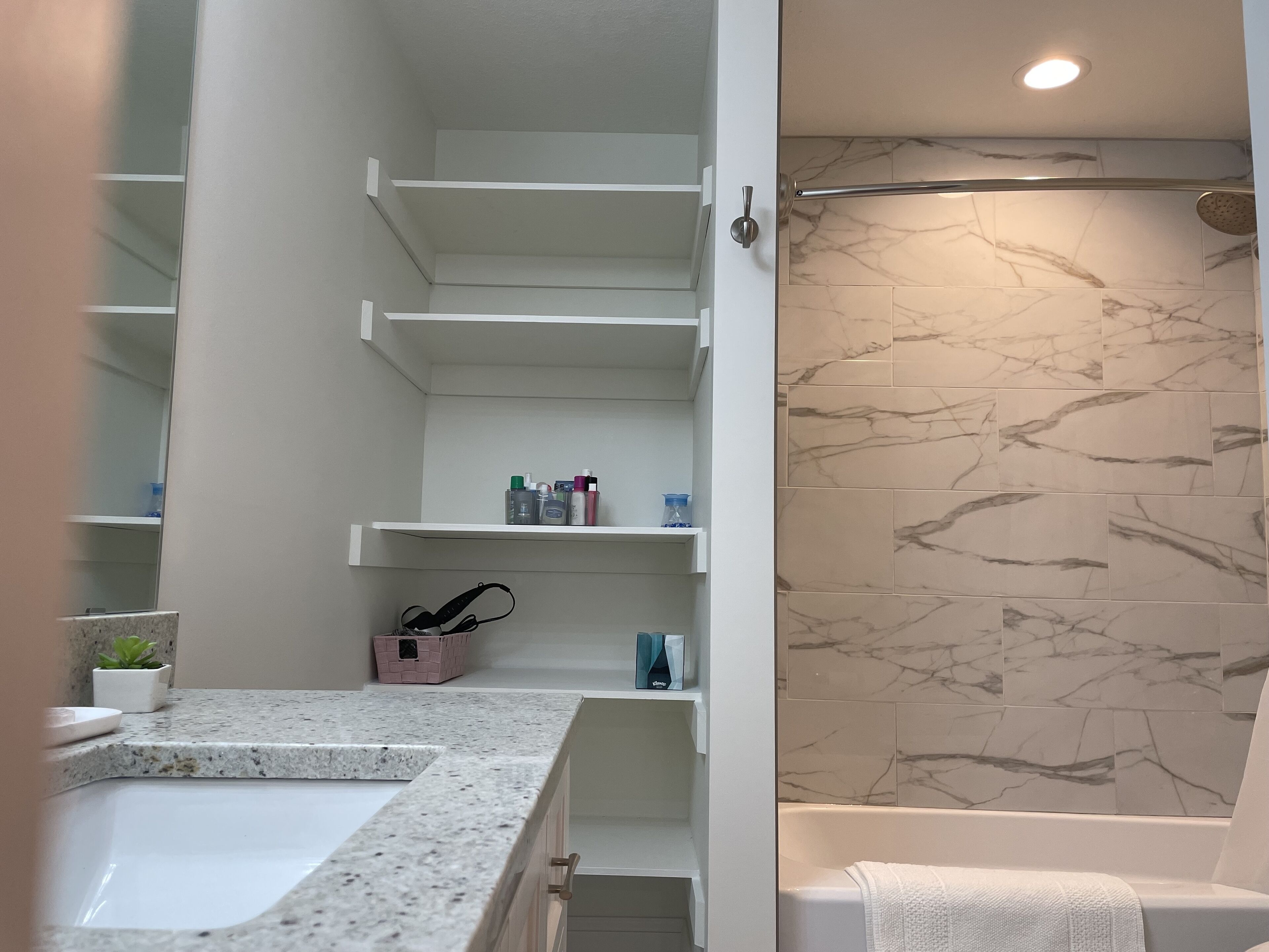 Casa de banho com parede de chuveiro em mármore, lavatório branco e prateleiras embutidas com artigos de higiene pessoal.
