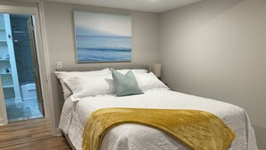 A bedroom with a bed, white bedding, a yellow throw blanket, a blue pillow, and a painting of the sea on the wall.