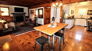 A spacious living area with a wooden floor, a fireplace, a dining table with stools, and a kitchen in the background. - Great Home With Large Yard 300 Yards To Sheep's Pond (2277) (Brewster)