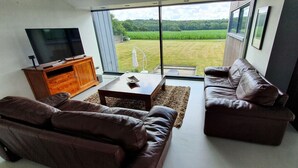 A living room with a large TV, leather sofas, a wooden cabinet, and a view of a green field through large windows. - VILLA COMBINES MODERNITY AND STONES. IDEAL FOR FAMILY OR WORK (LIFFRE)